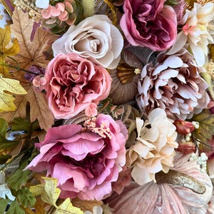 May include: A close-up of a fall-themed wreath with a variety of artificial flowers and leaves. The wreath features large pink peonies, white roses, and light-coloured dahlias. Gold and brown pumpkins are also incorporated into the design.