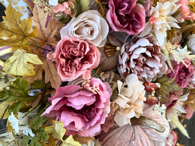 May include: A close-up of a fall-themed wreath with a variety of artificial flowers and leaves. The wreath features large pink peonies, white roses, and light-coloured dahlias. Gold and brown pumpkins are also incorporated into the design.