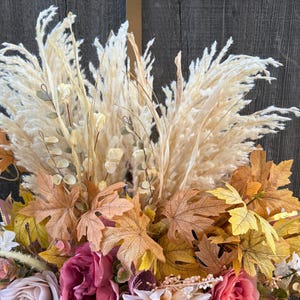 May include: A close-up of a fall-themed floral arrangement. The arrangement features large, cream-coloured pampas grass plumes, surrounded by faux fall leaves in shades of brown, yellow, and orange. Pink and cream-coloured roses are also visible.