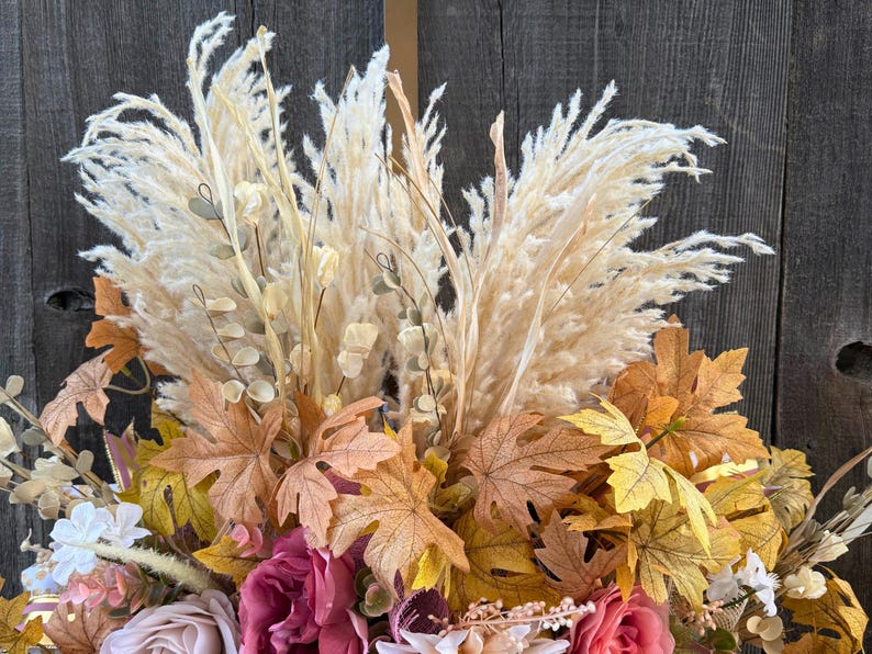 May include: A close-up of a fall-themed floral arrangement. The arrangement features large, cream-coloured pampas grass plumes, surrounded by faux fall leaves in shades of brown, yellow, and orange. Pink and cream-coloured roses are also visible.