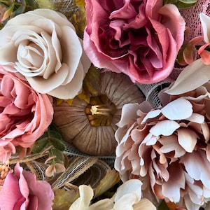May include: Close-up of a floral wreath with artificial flowers in pink, cream, and peach hues. The wreath includes roses, peonies, and a small pumpkin, creating a warm, autumnal theme.
