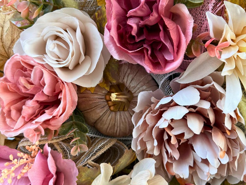 May include: Close-up of a floral wreath with artificial flowers in pink, cream, and peach hues. The wreath includes roses, peonies, and a small pumpkin, creating a warm, autumnal theme.