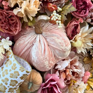 May include: A seasonal wreath with a plush, pale pink pumpkin at its heart. The wreath is decorated with an array of flowers in pink, peach, and cream hues, and includes ribbon with a gold leopard print design.