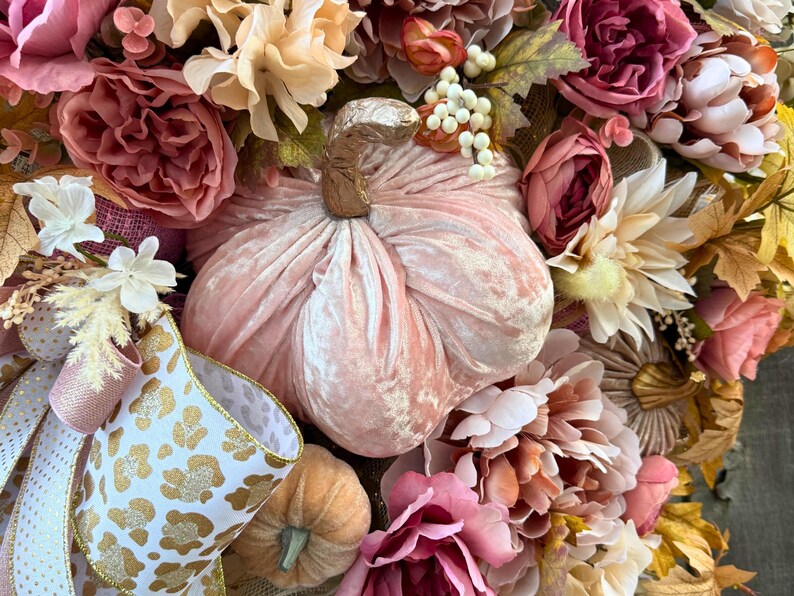 May include: A seasonal wreath with a plush, pale pink pumpkin at its heart. The wreath is decorated with an array of flowers in pink, peach, and cream hues, and includes ribbon with a gold leopard print design.