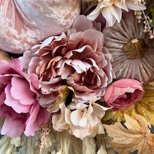 May include: A close-up of a fall-themed wreath with a variety of faux flowers and pumpkins. The wreath features large pink peonies, smaller pink buds, and tan and brown pumpkins. The colour palette includes shades of pink, tan, and brown.