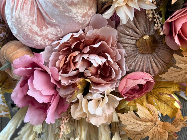 May include: A close-up of a fall-themed wreath with a variety of faux flowers and pumpkins. The wreath features large pink peonies, smaller pink buds, and tan and brown pumpkins. The colour palette includes shades of pink, tan, and brown.