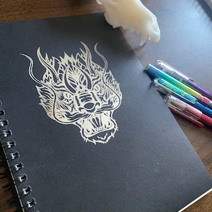 May include: A black notebook with a white dragon illustration on the cover. The notebook has a spiral binding and is lying on a wooden surface.