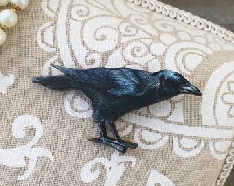 Puede incluir: Broche de cuervo negro, posado sobre una tela estampada. El ave presenta detalles de plumas y un pico afilado. Un collar de perlas con detalles rojos y morados es visible. Un accesorio decorativo para ropa o bolsos.