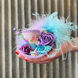May include: A miniature top hat fascinator featuring a lavender floral print base, adorned with purple and teal fabric roses, light blue feathers, and a small decorative teapot charm. The brim is trimmed with lavender braided cord.