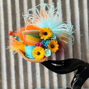 May include: A whimsical top hat with a straw brim, adorned with yellow sunflowers, blue and pink flowers, and colorful feathers. The hat features a striped fabric band and is accented with decorative beads.
