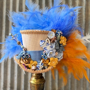 May include: A miniature top hat adorned with bright blue and orange feathers, featuring a beige textured crown and a blue brim. It is decorated with artificial flowers in shades of yellow, blue, and gray, a miniature teapot, a teacup, and a small spoon.