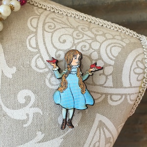 May include: A wooden pin of a girl in a blue and white gingham dress holding red shoes in each hand.
