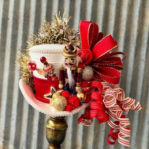 Op de afbeelding: Een feestelijke kerstversiering met een rood-witte hoed versierd met een houten notenkraker, gouden ornamenten en een grote rode strik. De hoed is versierd met gouden slingers en kralen, wat een grillige vakantieweergave creëert.