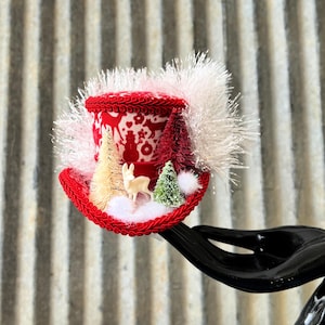 May include: A miniature red and white top hat with a winter holiday theme. The hat features a red brim and a white and red patterned cylinder with a winter scene. It is decorated with miniature trees, a deer, and white fluffy accents.