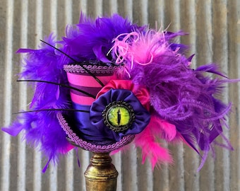 Pode incluir: Uma cartola com listras horizontais roxas e rosas, adornada com um arranjo denso de penas roxas e rosas vibrantes. Uma roseta de tecido roxo escuro fica na frente, centralizada com um cabochão realista de olho de gato amarelo e preto.