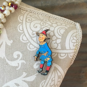 May include: A colorful pin of the Scarecrow from The Wizard of Oz. The pin features the Scarecrow in his classic outfit: a blue outfit, red boots, and a black hat with a red brim. The pin is displayed on a patterned fabric.