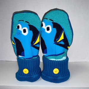 May include: A pair of blue and green knit gloves with a cartoon fish design. The gloves have a yellow button on the cuff.