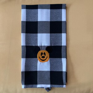 Puede incluir: Un pa&ntilde;o de cocina a cuadros blancos y negros con un rostro bordado de Mickey Mouse en el centro. La cara de Mickey Mouse es naranja con ojos negros y una sonrisa negra. La toalla est&aacute; doblada y tiene un lazo en la parte inferior.