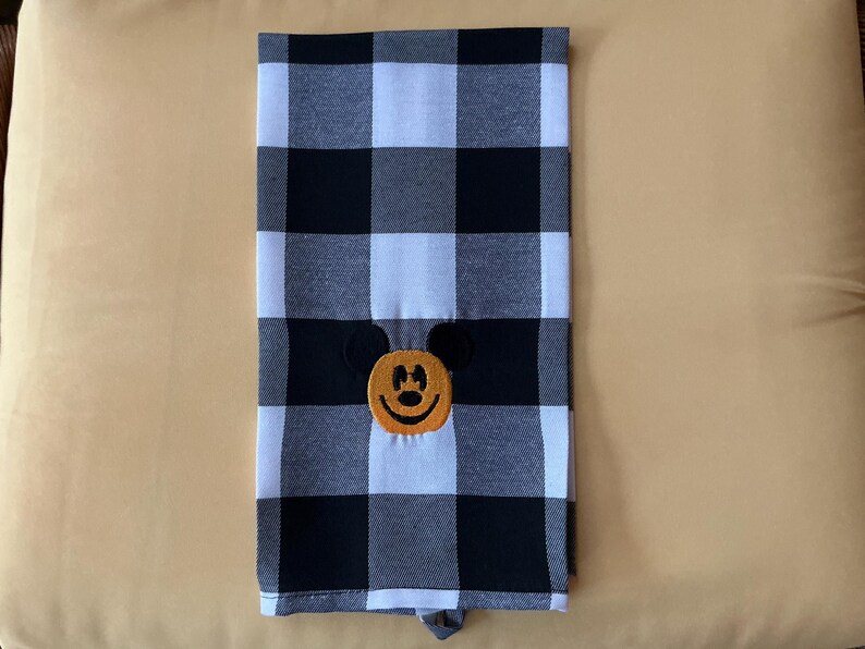 Puede incluir: Un pa&ntilde;o de cocina a cuadros blancos y negros con un rostro bordado de Mickey Mouse en el centro. La cara de Mickey Mouse es naranja con ojos negros y una sonrisa negra. La toalla est&aacute; doblada y tiene un lazo en la parte inferior.