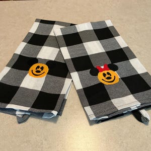 Puede incluir: Dos pa&ntilde;os de cocina a cuadros blancos y negros. Una toalla presenta una cara de Mickey Mouse bordada, y la otra una cara de Minnie Mouse bordada con un lazo rojo. Las toallas est&aacute;n dobladas y sobre una superficie clara.