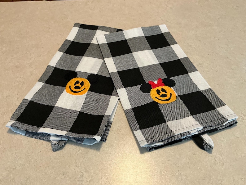 Puede incluir: Dos pa&ntilde;os de cocina a cuadros blancos y negros. Una toalla presenta una cara de Mickey Mouse bordada, y la otra una cara de Minnie Mouse bordada con un lazo rojo. Las toallas est&aacute;n dobladas y sobre una superficie clara.