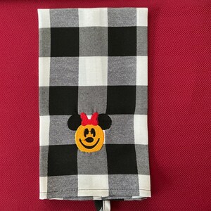 Puede incluir: Un pa&ntilde;o de cocina a cuadros blancos y negros con un rostro de Minnie Mouse bordado. La cara de Minnie Mouse es amarilla con orejas negras y un lazo rojo. La toalla est&aacute; doblada y tiene una etiqueta blanca.
