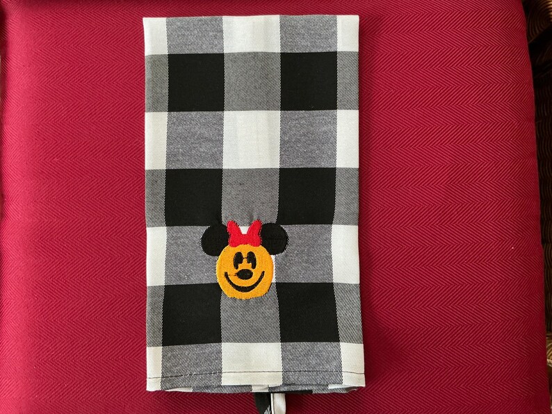 Puede incluir: Un pa&ntilde;o de cocina a cuadros blancos y negros con un rostro de Minnie Mouse bordado. La cara de Minnie Mouse es amarilla con orejas negras y un lazo rojo. La toalla est&aacute; doblada y tiene una etiqueta blanca.