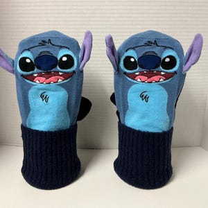 May include: A pair of blue mittens featuring the character Stitch from the Disney movie Lilo & Stitch. The mittens have a blue body, light blue belly, purple ears, and a navy blue ribbed cuff. The character's face is embroidered on the front.