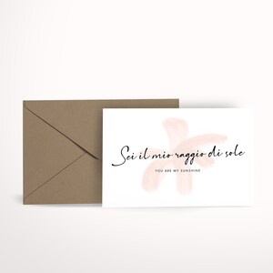 Può includere: Un biglietto bianco con un disegno ad acquerello rosa e il testo "Sei il mio raggio di sole" in corsivo nero. Sotto il testo c'è la frase "You are my sunshine" in un carattere nero più piccolo.