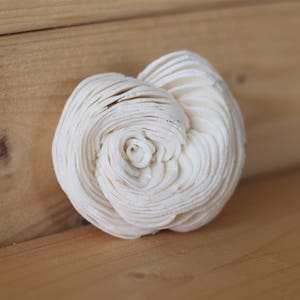 May include: A cream-colored, spiral-shaped decorative flower made of layered material. The flower is centered on a light brown wooden surface, with the wood grain visible. The flower's design resembles a rose.