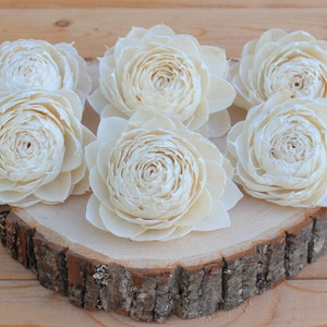 May include: Six ivory-colored, handcrafted sola wood flowers arranged on a rustic wooden slice. The flowers have multiple layers of petals, creating a full, open bloom. The wood slice has a natural bark edge.