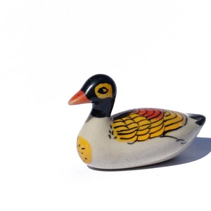 May include: A small ceramic duck figurine with a white body, black head, and yellow and red painted details. The duck is lying on its belly with its head turned to the side.