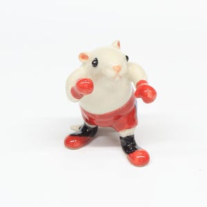Puede incluir: Una figurita de cerámica blanca de un ratón con guantes de boxeo rojos y pantalones cortos de boxeo rojos y negros.