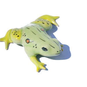 Könnte beinhalten: Eine grüne Keramik-Froschfigur mit gelben Akzenten und schwarzen und orangefarbenen Flecken. Der Frosch hat eine strukturierte Oberfläche und einen verspielten Ausdruck.