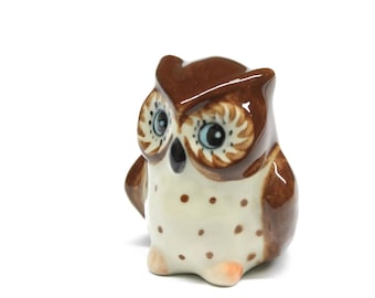 Könnte beinhalten: Eine kleine Keramik-Eulenfigur, die in Braun und Weiß bemalt ist. Die Eule hat große, runde Augen mit blauen Pupillen und einen weißen Schnabel.