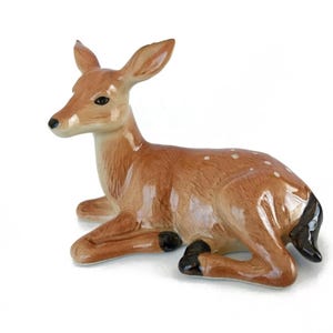 May include: A ceramic fawn figurine with brown and white spots, lying on its belly with its head turned to the side. The fawn has black hooves and a black tail.