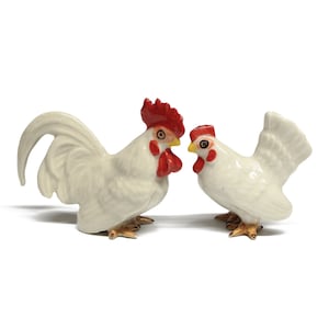 Op de afbeelding: Twee witte keramische beeldjes van hanen. De haan links heeft een rode kam en een rode keellap. De haan rechts heeft een rode kam en een rode keellap en kijkt naar links.