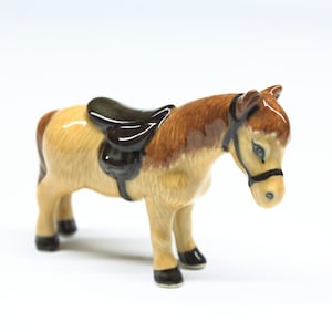 May include: A small ceramic figurine of a brown and tan horse with a black saddle. The horse has a black bridle and is standing on a white surface.