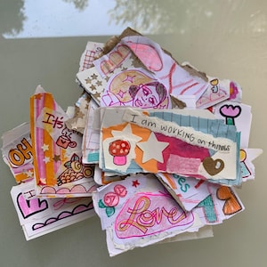 May include: A pile of colorful, torn paper scraps with hand-drawn illustrations and text. The drawings include a mushroom, stars, a heart, and the word "Love". The text fragments read "I am working on things" and "It's".