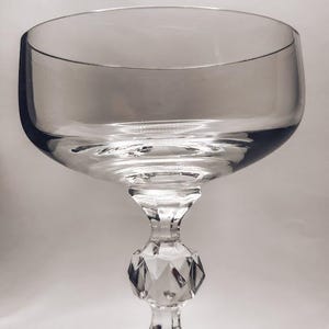 May include: A clear, stemmed champagne coupe glass. The bowl is wide and shallow, supported by a faceted stem and a circular base. The glass is transparent, reflecting light.