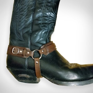 May include: A pair of black leather cowboy boots with a brown leather strap around the ankle. The strap has a silver buckle and a metal ring.