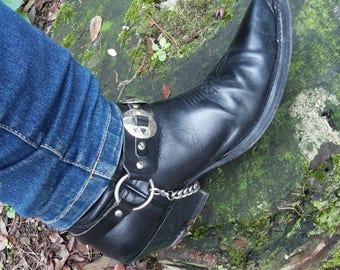 May include: A black leather cowboy boot with a silver buckle and chain detail. The boot is worn with blue jeans.