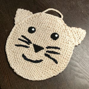 Può includere: Un sottopentola beige a forma di gatto, lavorato all'uncinetto con tratti del viso ricamati in nero, tra cui occhi, baffi e naso. Il sottopentola ha un anello per appenderlo ed è progettato per proteggere le superfici dal calore.