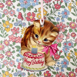 May include: A vintage-style birthday card featuring a cartoon kitten holding a pink and white birthday cake with a single lit candle. The card has a "Happy Birthday" message on the cake and is set against a background of a floral pattern.