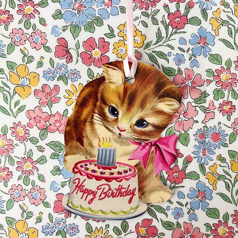 May include: A vintage-style birthday card featuring a cartoon kitten holding a pink and white birthday cake with a single lit candle. The card has a "Happy Birthday" message on the cake and is set against a background of a floral pattern.