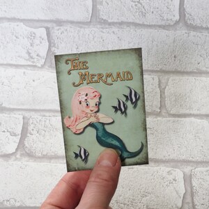 May include: A vintage-style illustration of a mermaid with pink hair and a green tail. The illustration is on a light green background with the text "The Mermaid" in gold lettering. There are black and white fish swimming around the mermaid.