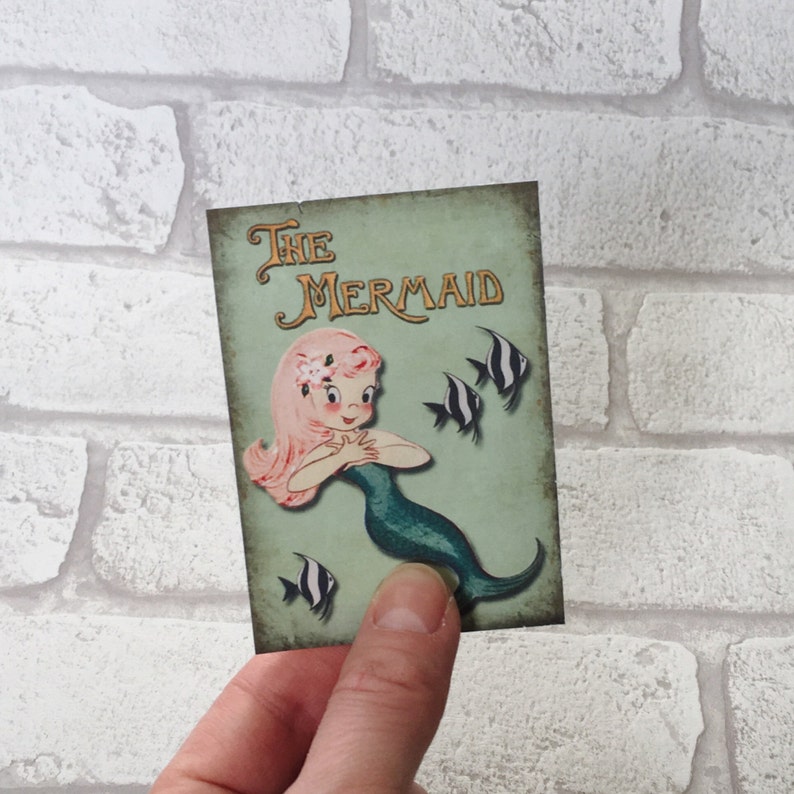 May include: A vintage-style illustration of a mermaid with pink hair and a green tail. The illustration is on a light green background with the text "The Mermaid" in gold lettering. There are black and white fish swimming around the mermaid.