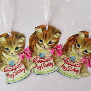 May include: Three brown and white kitten gift tags with pink ribbons and a birthday cake with a single lit candle. Each tag says "Happy Birthday".