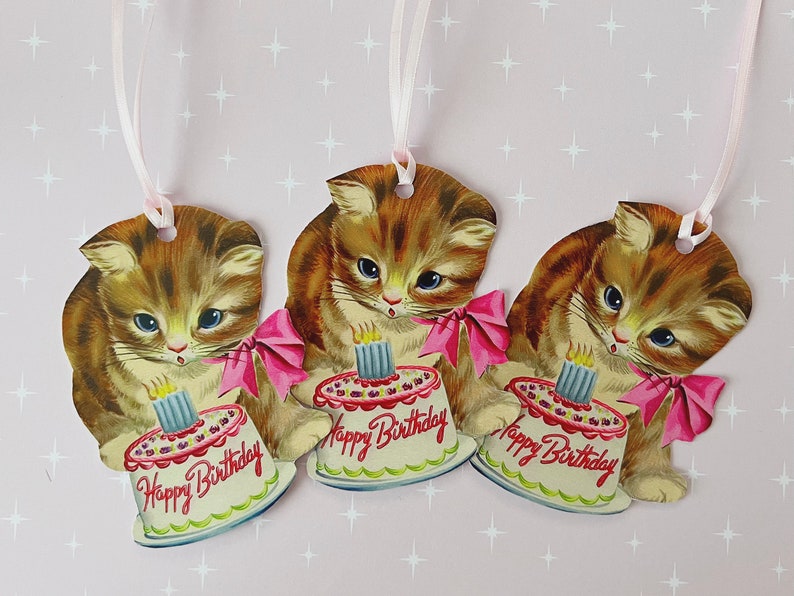 May include: Three brown and white kitten gift tags with pink ribbons and a birthday cake with a single lit candle. Each tag says "Happy Birthday".