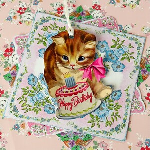 May include: A vintage birthday card featuring a cartoon cat holding a cake with the words "Happy Birthday" on it. The card is decorated with blue and pink flowers.